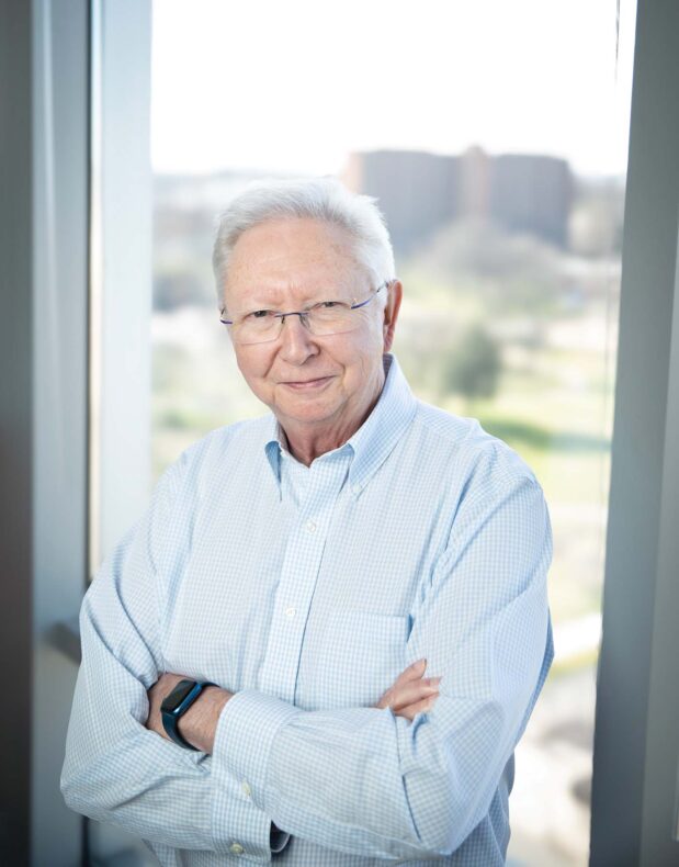 Frank Harrell Jr, PhD, is a long-time biostatistician at VUMC. He is pictured in a light blue checked button down with arms folded standing against a window. 