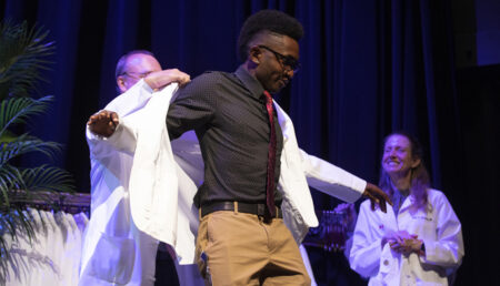 Amy Fleming, MD, looks on as Jeff Balser, MD, PhD, helps Dominic Okonkwo into his white coat at last week’s ceremony.